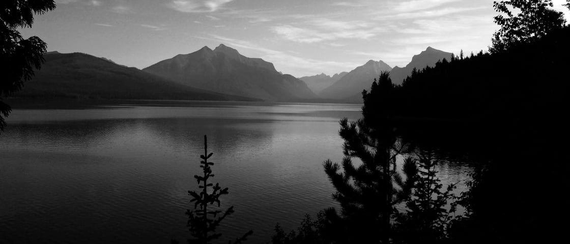 Black and white photograph of a mountain lake in Glacier National Park. Mountains are in the background and trees surround the lake in the foreground.