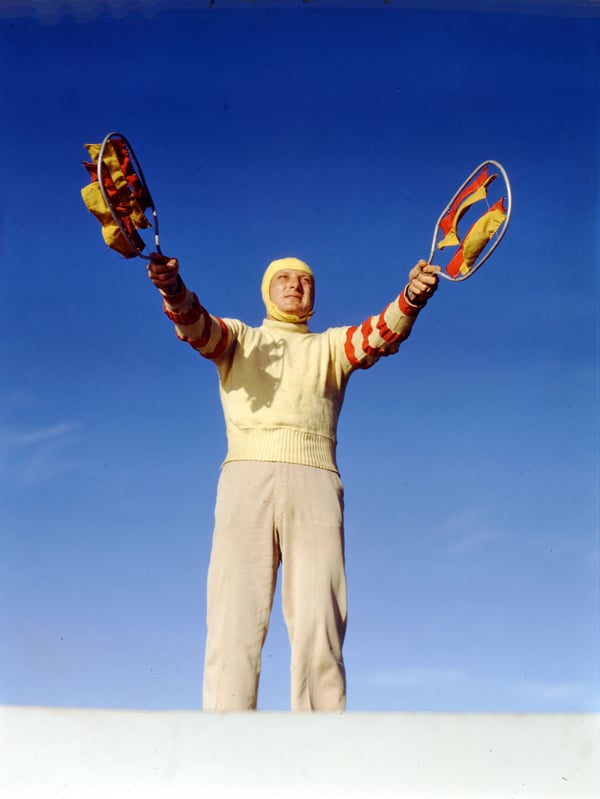 Photograph of a landing safety officer guiding in a plane with a flag in each hand. A bright blue sky is behind him.