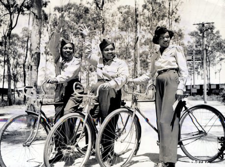 Black and white photograph of 3 young women in uniforms standing next to their bicycles. They are smiling and looking off camera