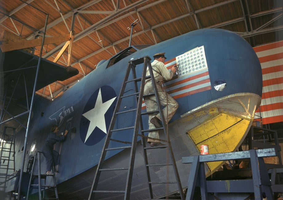 Color photograph of two service members standing on ladders and painting the sides of an ocean aircraft.