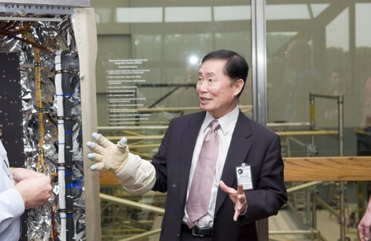 Actor George Takei at Goddard Space Flight center with a glove on his hand talking to someone off camera.