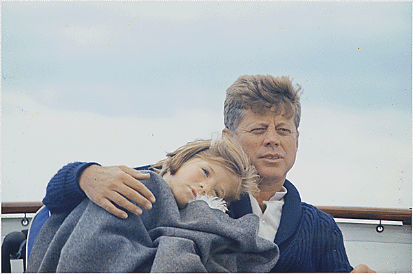 Color photograph of John F. Kennedy on a boat sitting next to his daughter Caroline. She is wrapped in a blanket and leans her head on his shoulder while he wraps his arm around her.
