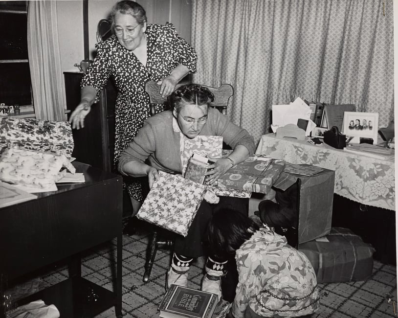 Black and white photograph of two women wrapping presents while a small child crouches near them on the floor