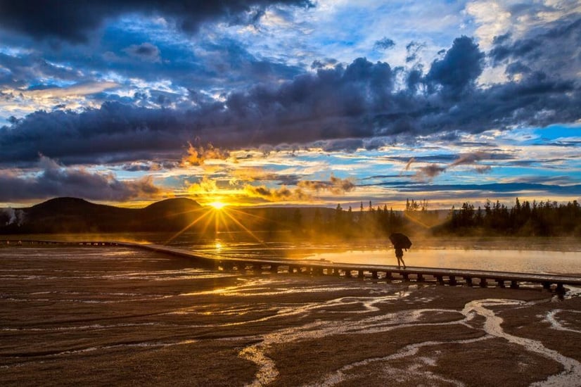 Color photograph of a person walking over a bridge with a dramatic sunrise in front of them. The cloudy with multiple shades of blue, while the water and land is a golden color 