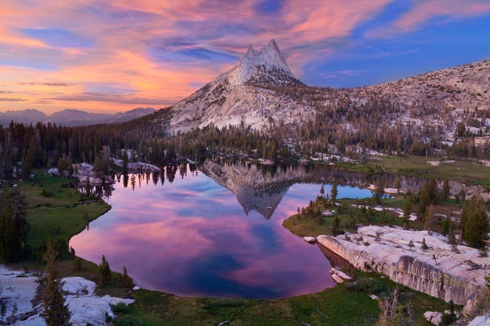 Color photograph of a snow capped mountain in Yosemite National Park. The sky behind the mountain is blue and orange, which is reflected in a body of water at the base of the mountain. 