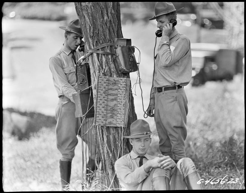 Black and white photograph of 3 soldiers standing by a tree trunk. One sits on the ground facing the camera, while the other two use a telephone attached to equipment on the tree.