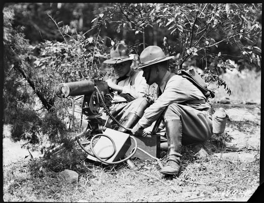 Black and white photograph of two soldiers crouched on the ground by tree branches. They have guns and communication devices.