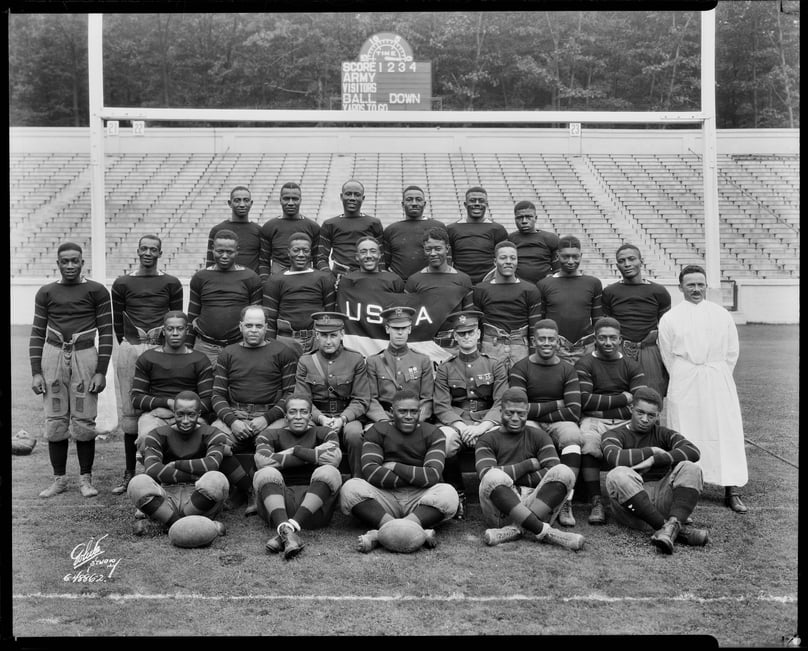 Black and white photograph of a team of football players. The players are in uniform with a goal post and stadium behind them. 