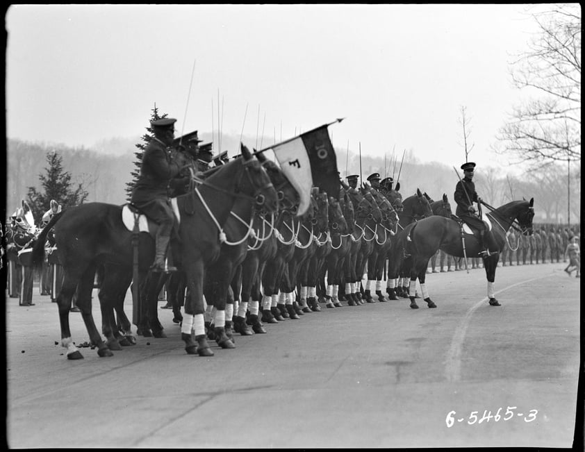 Black and white photograph of a parade of soldiers on horseback. One soldier carries a two colored flag reading 