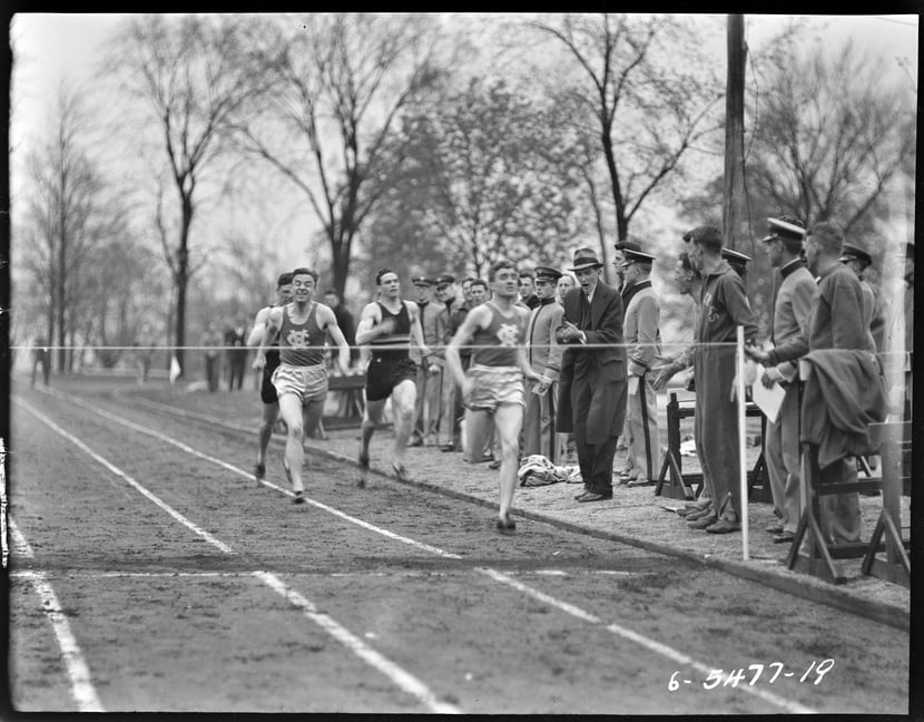 Black and white photograph of a track race. Several runners approach the finish line, while spectators cheer on from the sidelines.