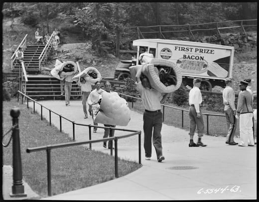 Black and white photograph of a group of people carrying personal items, including mattresses, down a walkway. A bacon truck appears in the background.