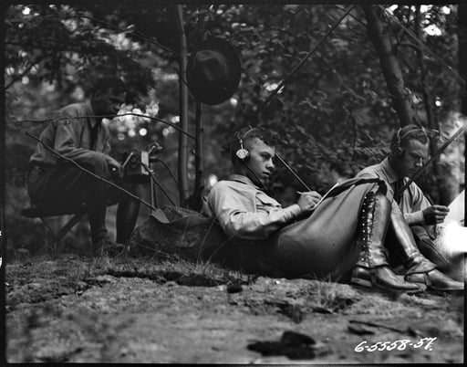 Black and white photograph of a soldier sitting on the ground, leaning against a tree.  He is wearing headphones and writing something in his lap. A hat is hanging on a tree branch above him. 