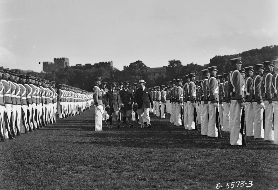 Black and white photograph of lines of soldiers standing at attention facing to the left. A group of men is walking in between the lines of soldiers. 