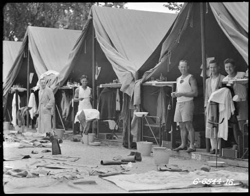 Black and white photograph of a group of camp tents. Soldiers stand at the entrance to the tents, smiling at the camera