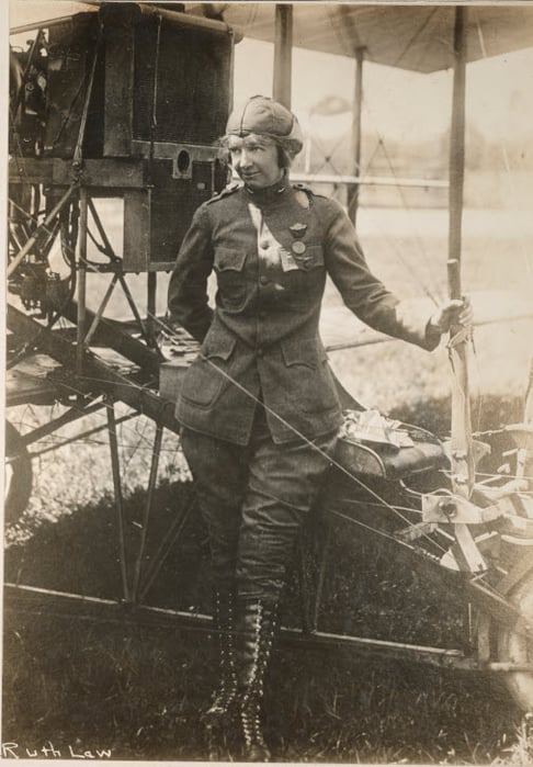 Black and white photograph of Ruth Law in flying uniform, standing next to a plane.