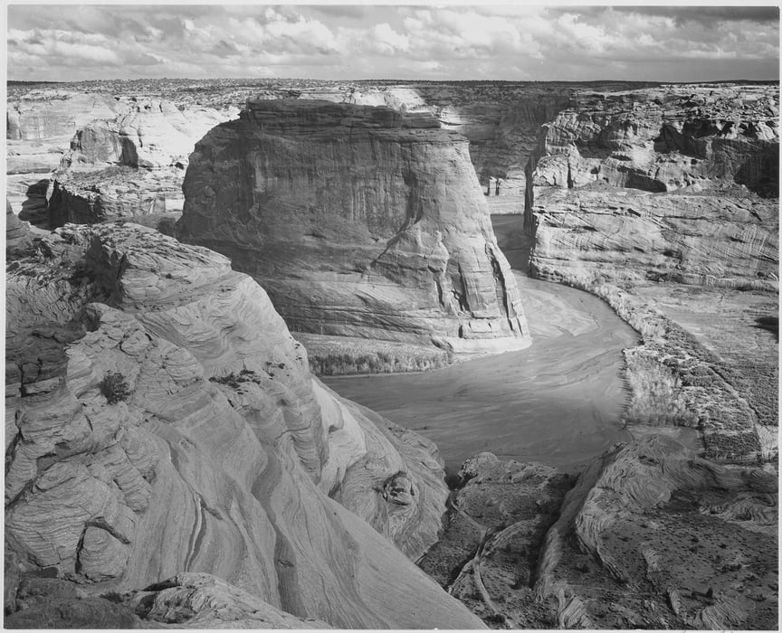 Black and white photograph of a canyon in Canyon de Chelly in Arizona. A river winds through the canyon around the mountain.
