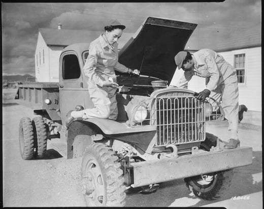 Black and white photograph of two women working on a car. The car's hood is open