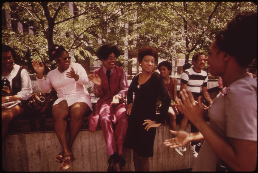 Photograph of a group of people in a park talking and laughing. Some are standing, some are sitting on a fountain ledge