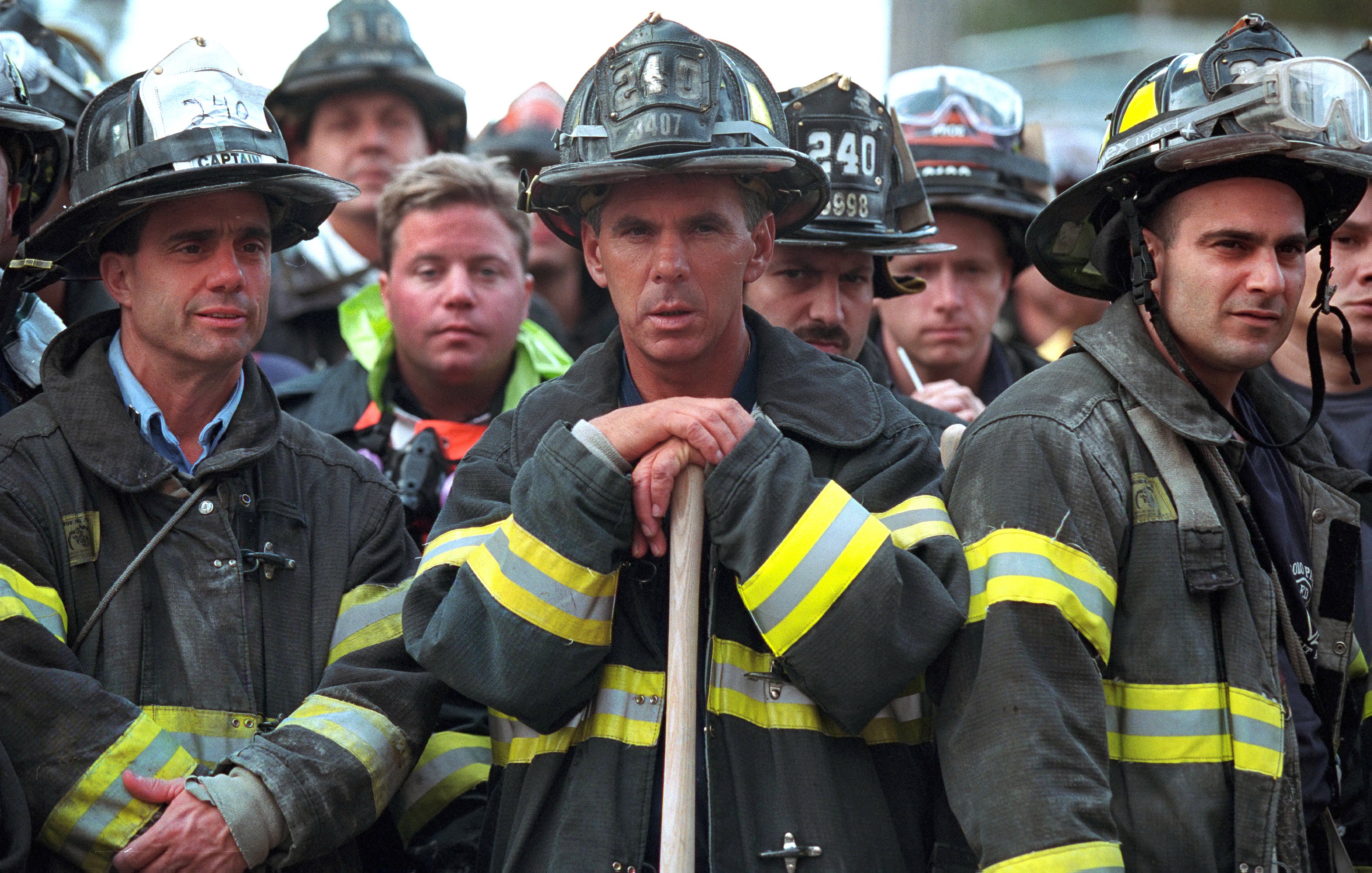 Group of firefighters in uniform in New York City stand together, looking on as President Bush surveys the area following the 9/11 terrorist attacks