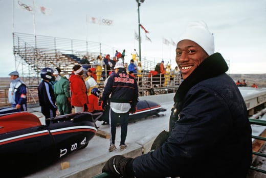 Bobsled team member looks back at the camera and smiles while other teammates and crew members are seen in the background.