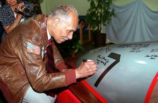 Photograph of a man in a brown leather jacket signing the outside of a plane with marker