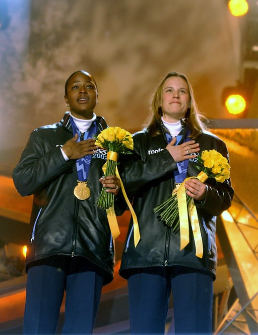 Photograph of Vonetta Flowers and Jill Bakken with their gold medals at the Olympic medal ceremony. Both women have their hands on their hearts and are holding flowers while singing the National Anthem.
