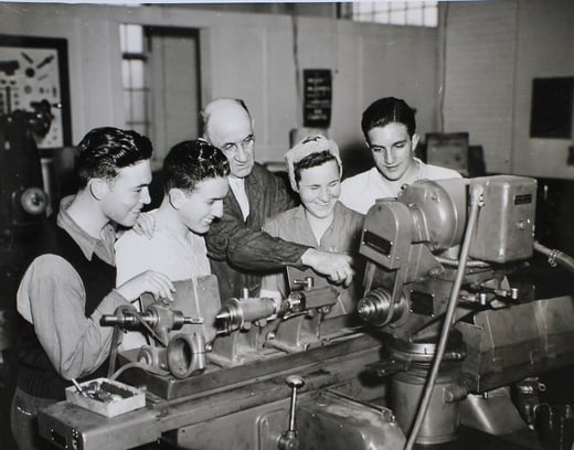 Black and white photograph of a group of young people receiving instruction in a workshop