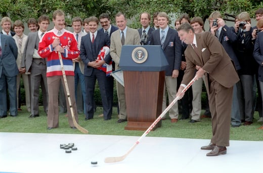 President Ronald Reagan holds a hockey stick and attempt to hit a puck while the U.S. Olympic Hockey team looks on.
