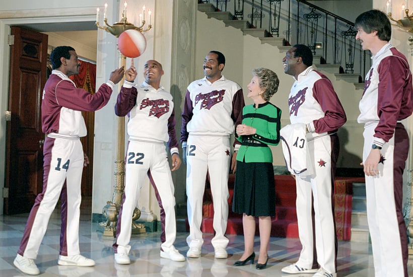 Photograph of Nancy Reagan standing among members of the Shooting Stars Basketball team. Two players spin a basketball on their index fingers while the rest of the group smiles and looks on.