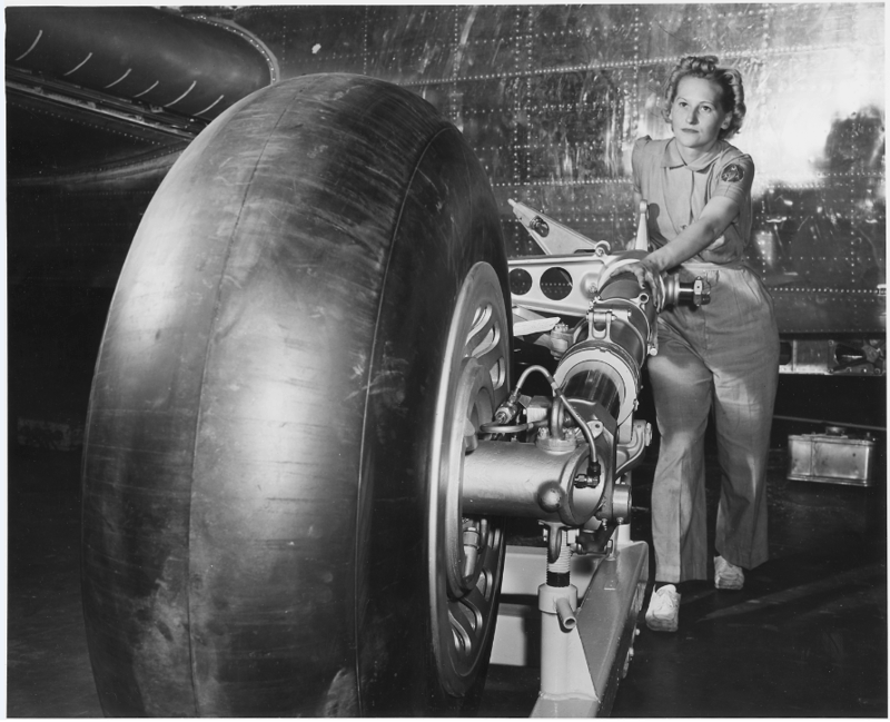 Black and white photograph of a woman rolling landing gear into place in a plant assembly line.