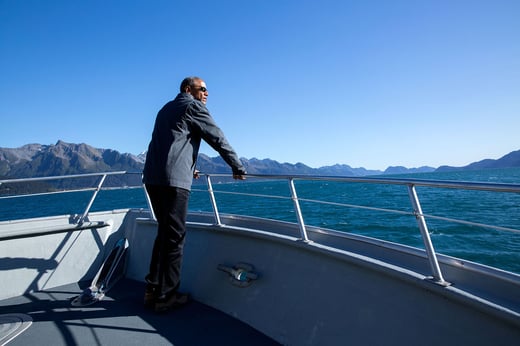 President Barack Obama stands at the railing of a ship in Kenai Fjords National Park. He looks out over the water at a mountain view.  The sky and water are clear blue.